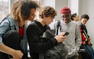five teenagers in a hallway looking at something on one the cell phone of one of the teenagers