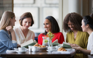 Group of five women seated at a table together looking at books and eating croissants.