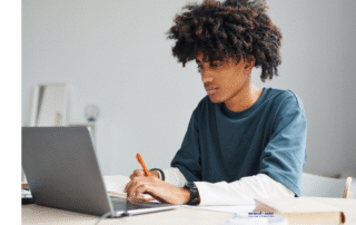 Teenage boy sitting at a desk working on a laptop and writing in a notebook