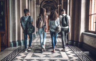 a group of four teenagers walk down a school hallway with backpacks on their shoulders
