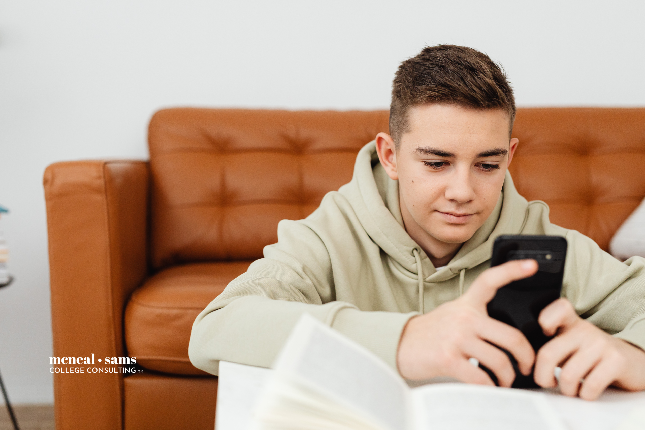 Teenage boy sits on floor in front of leather couch and scrolls on his phone.