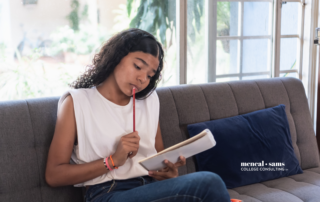 teenage girl sits on a grey couch looking thoughtfully at a notebook as she holds a pencil up to her mouth, pondering at the words on the page