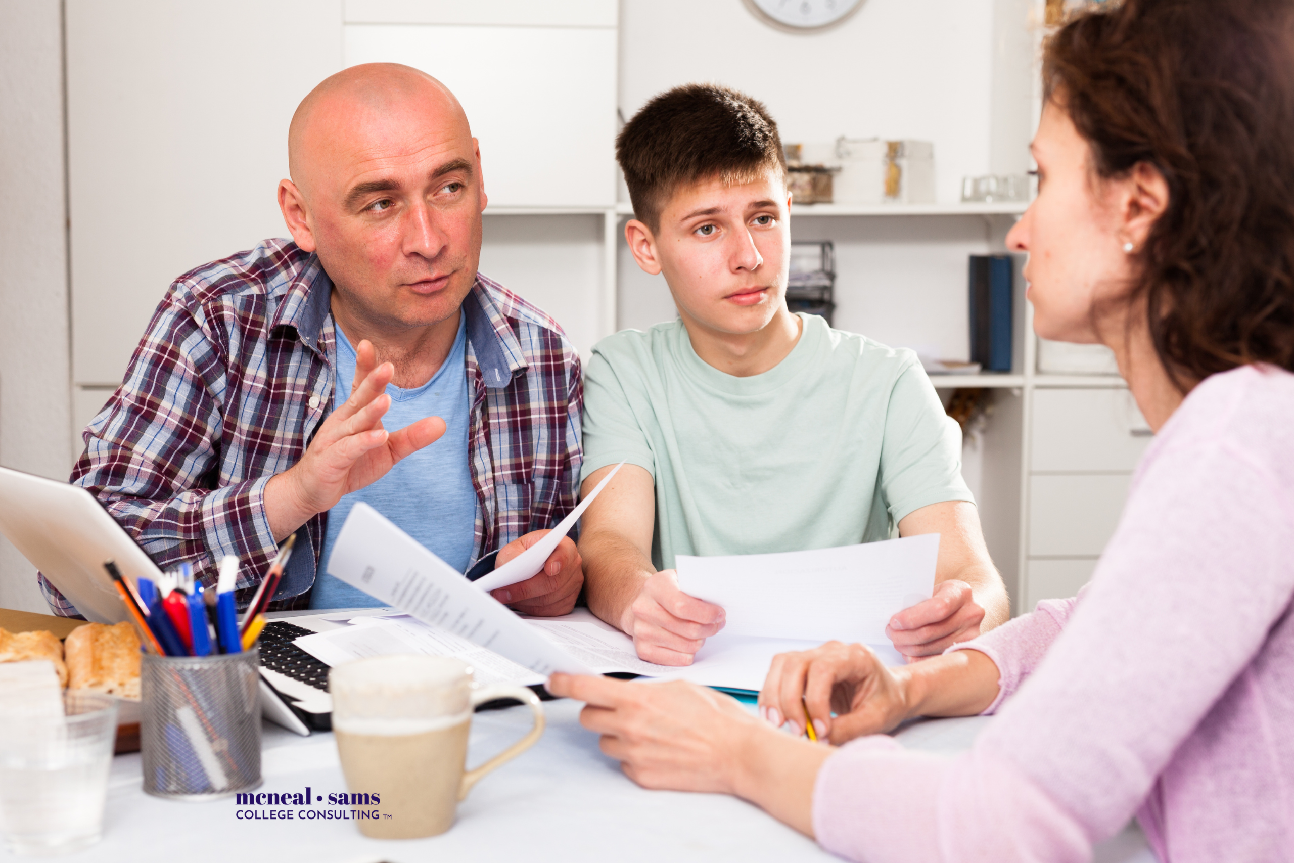 a man, woman, and a teenaged boy are sitting at a table talking as they look at papers.