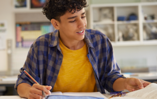 teenage boy is seated at a desk studying papers with a pencil in his hand