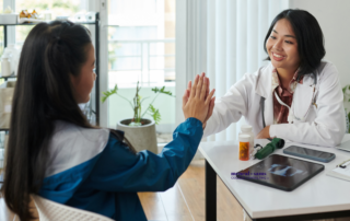 a teenage girl and her doctor high five while seated at a desk in an office