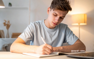A teenage boy sits at his desk, writing in a notebook.