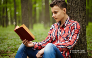 teenage boys sits against a tree in a forest reading a book
