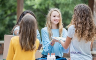 two teenage girls hand out notebooks and glue bottles to two other girls outdoors with trees behind them