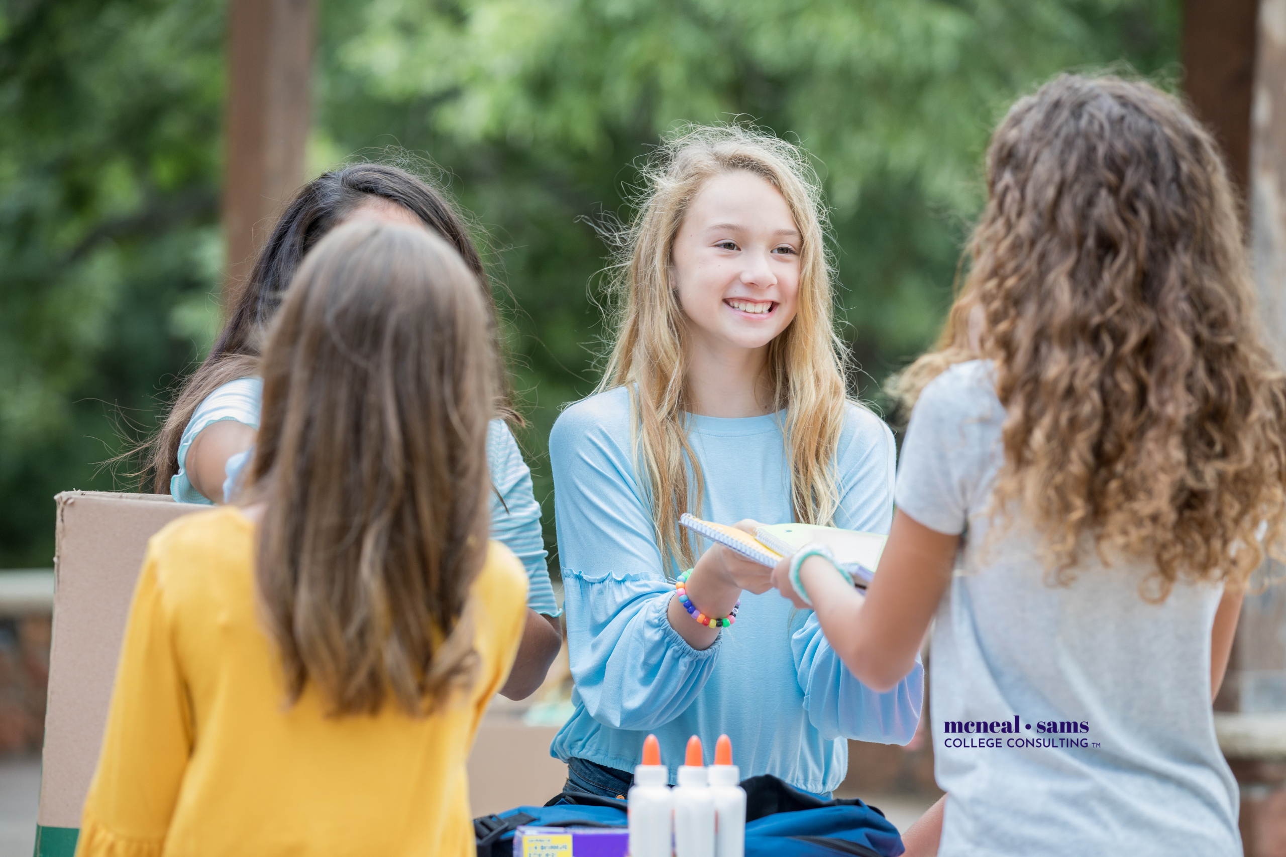 two teenage girls hand out notebooks and glue bottles to two other girls outdoors with trees behind them