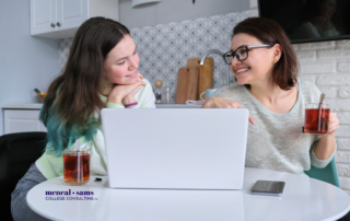Mother and teenage daughter sit at a table smiling at each other while looking at a laptop