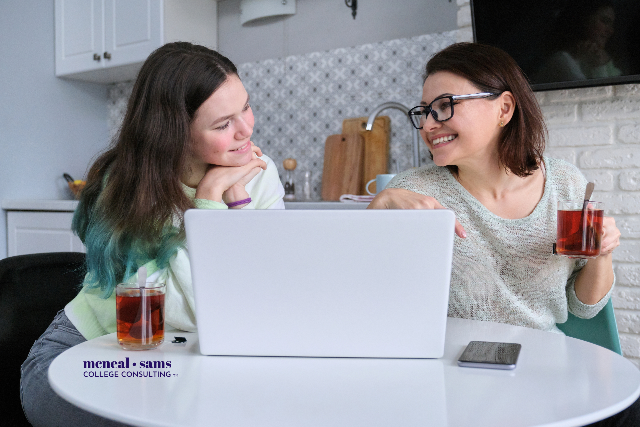 Mother and teenage daughter sit at a table smiling at each other while looking at a laptop