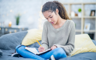 Teenage girl in a gray long sleeve top and jeans writes in her journal while sitting on her bed.