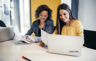 two teenage girls sit at a desk analyzing charts on paper and looking at a laptop screen.