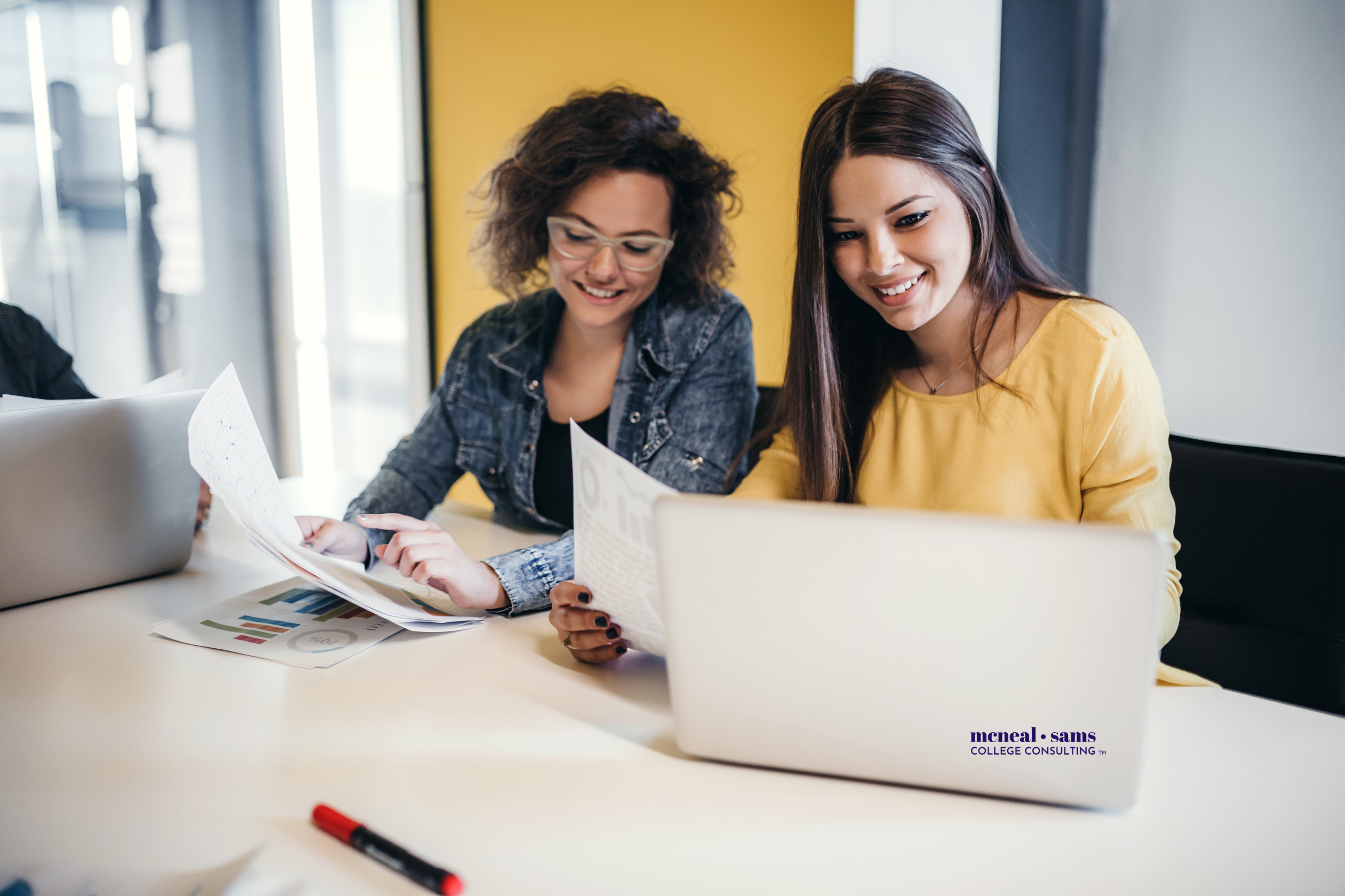 two teenage girls sit at a desk analyzing charts on paper and looking at a laptop screen.