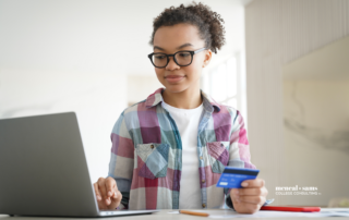 a young woman with glasses sitting at a desk enters a credit card number into her computer to make an online purchase
