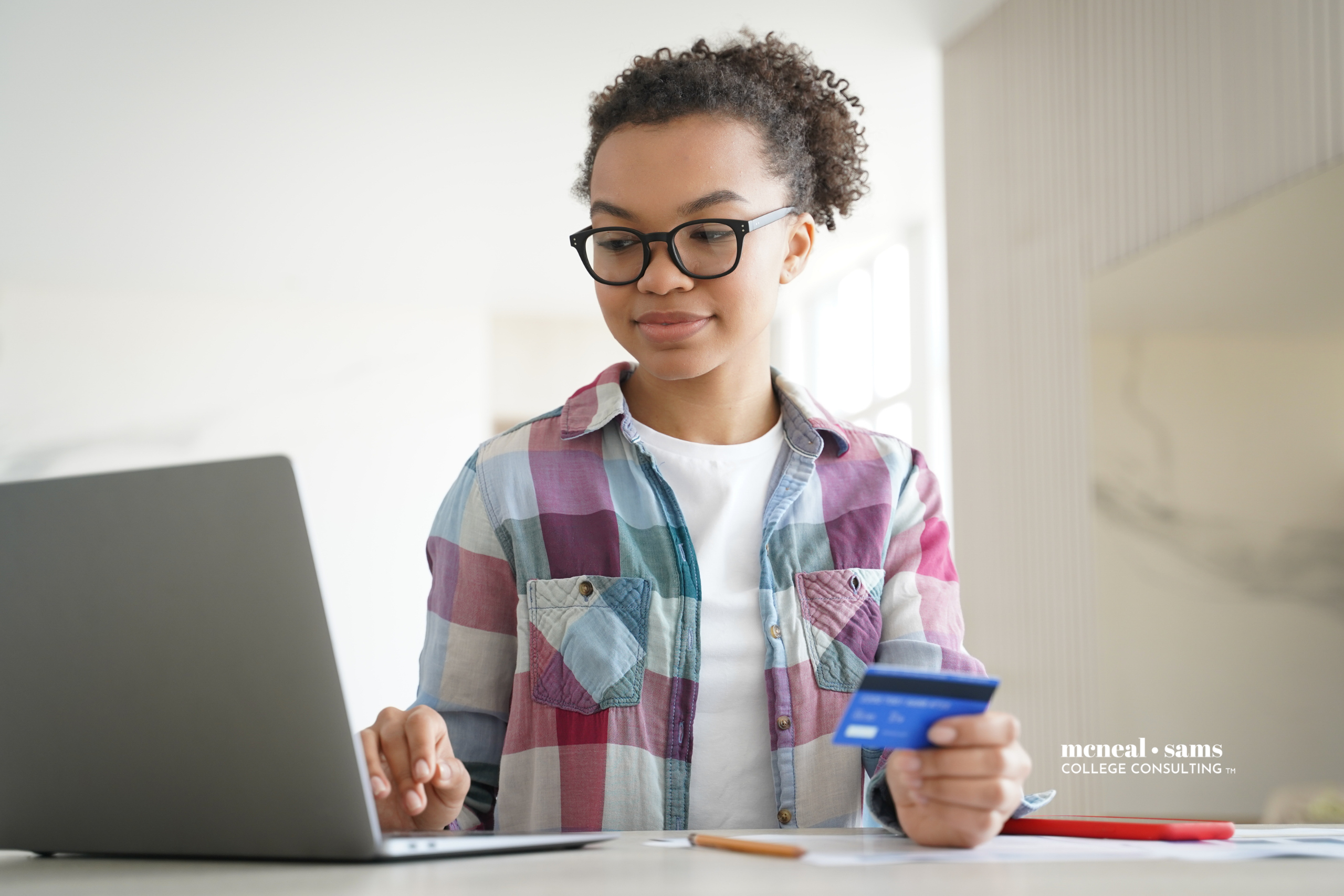 a young woman with glasses sitting at a desk enters a credit card number into her computer to make an online purchase