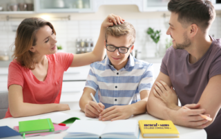 teenage boy sits with father and mother at a table while writing in a notebook