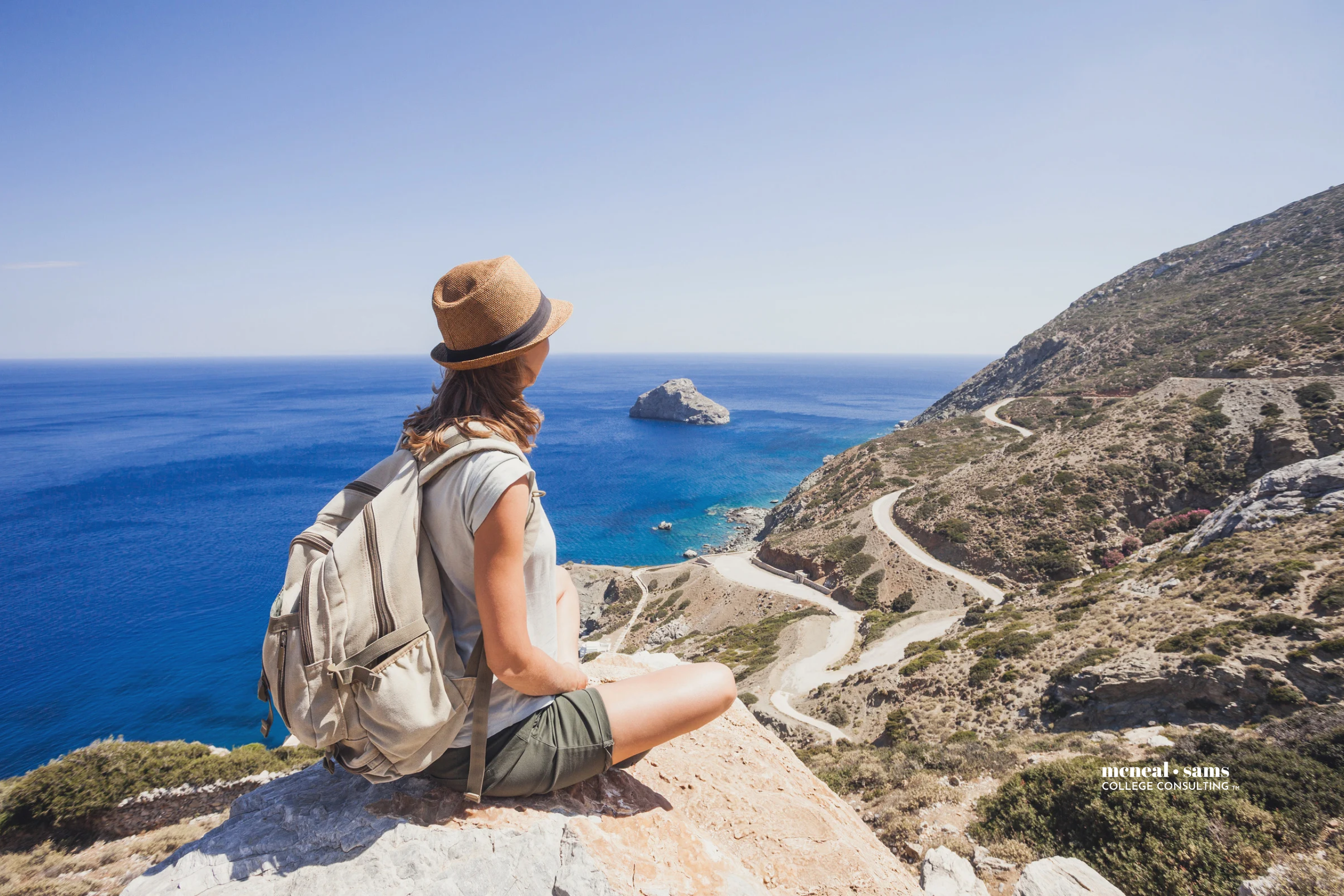 young woman wearing a backpack sits on the edge of a cliff overlooking the ocean while studying abroad